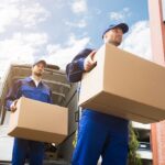 Close-up Of Two Delivery Men Carrying Cardboard Box