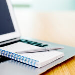 Laptop with pen and spiral notebook on desk.